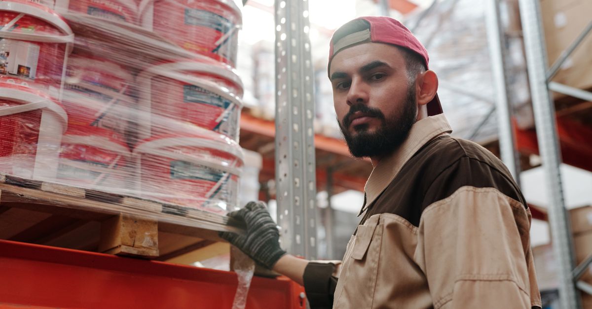 Portrait of a male warehouse worker standing next to stacked paint buckets indoors.
