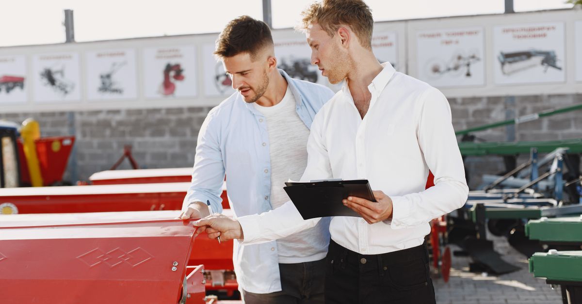 Two men discussing agricultural equipment purchase at a dealership, one holding a clipboard outside.