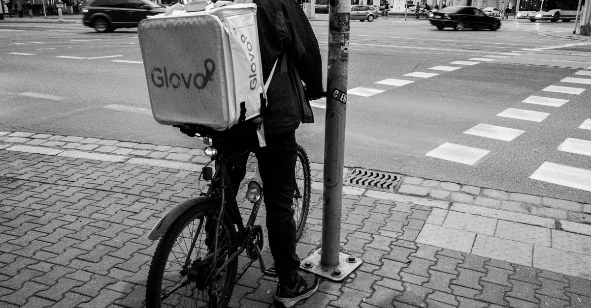 A delivery cyclist with a Glovo bag waits by a city street. Urban logistics in action.