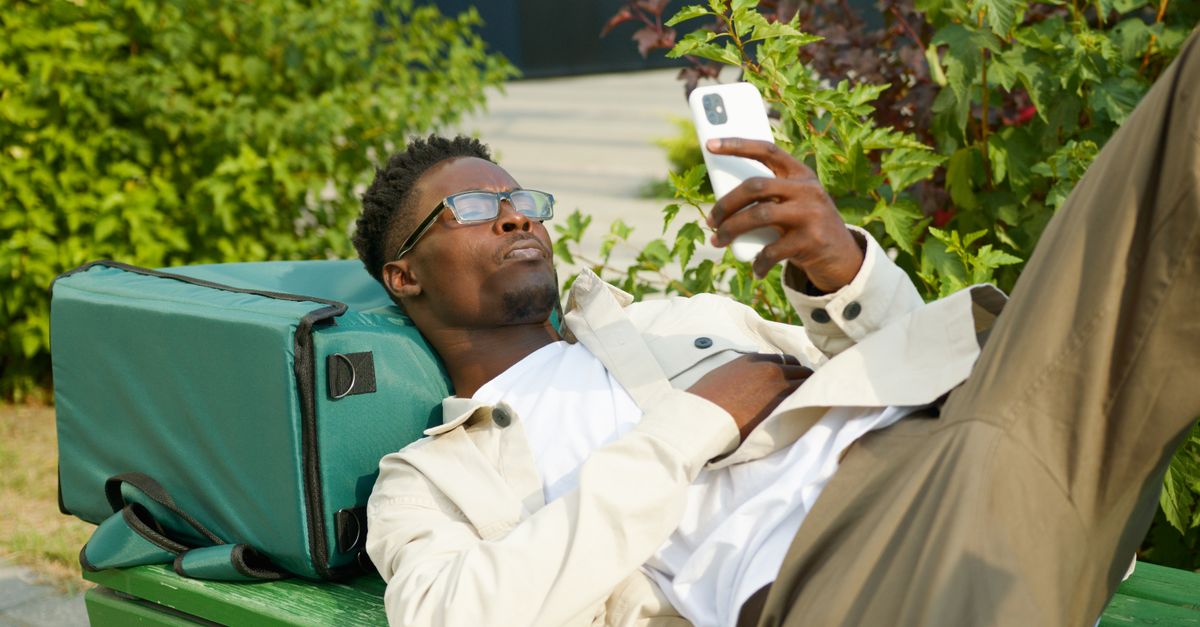 A delivery man lying on a bench outdoors, checking his smartphone beside his delivery bag.