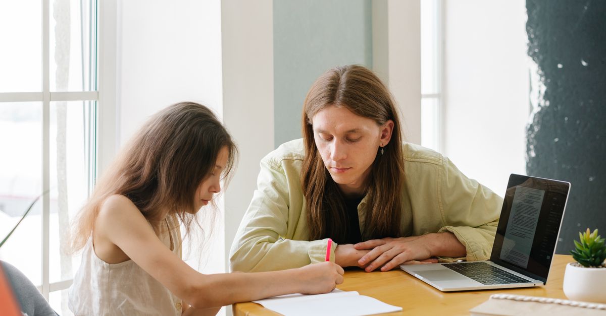 A father helps his daughter with homeschooling indoors, featuring a laptop and study materials.