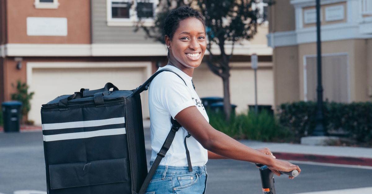 African American female courier delivering packages on a scooter with a thermal bag outdoors.