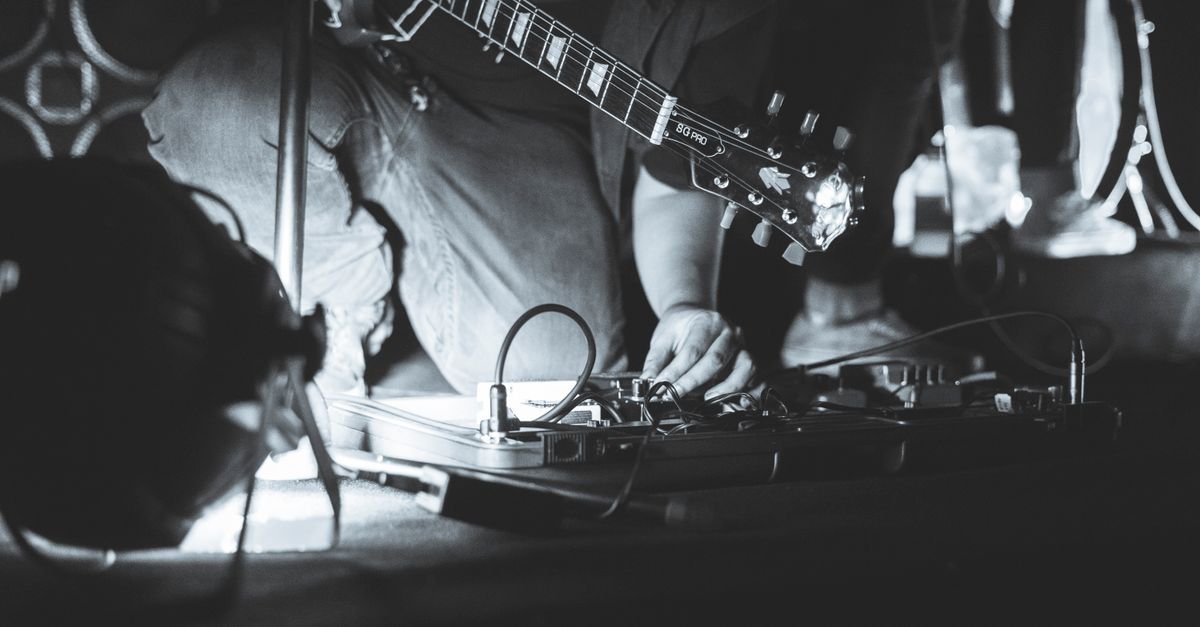 A musician adjusts guitar effects pedals on stage, captured in a candid black and white shot.