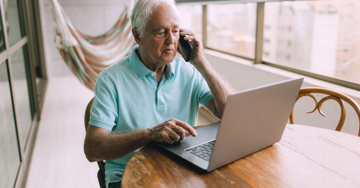 Elderly man multitasking with a phone call and laptop on a balcony.