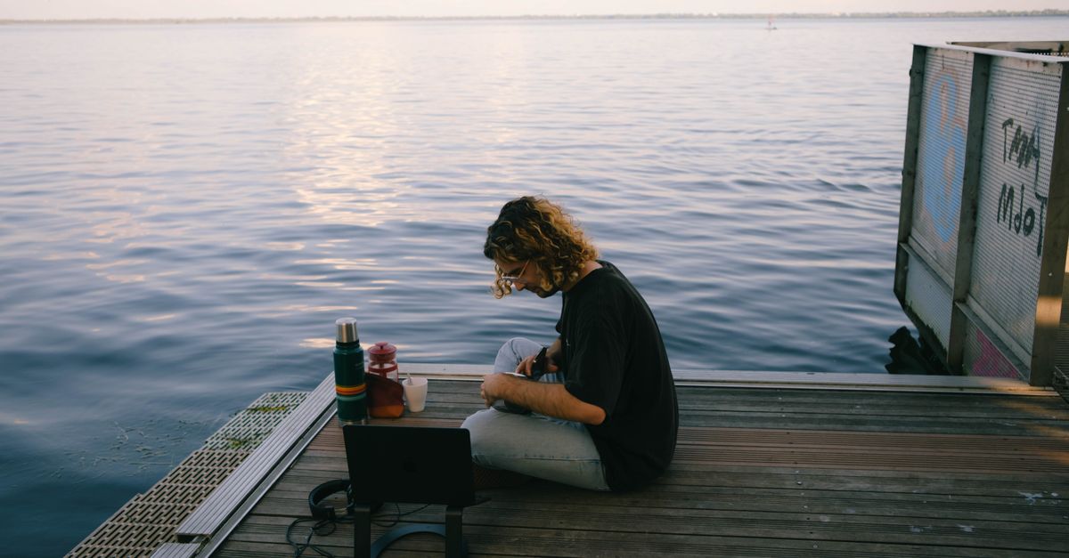 A man sits on a pier, immersed in nature, working on a laptop by the sea.