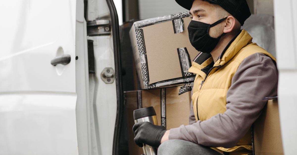 A courier wearing a face mask is seated in a van surrounded by packages, representing modern delivery services.