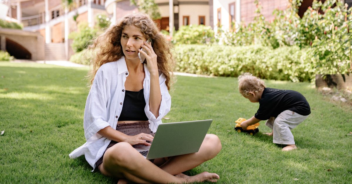 Woman multitasking on phone and laptop while child plays outdoors.
