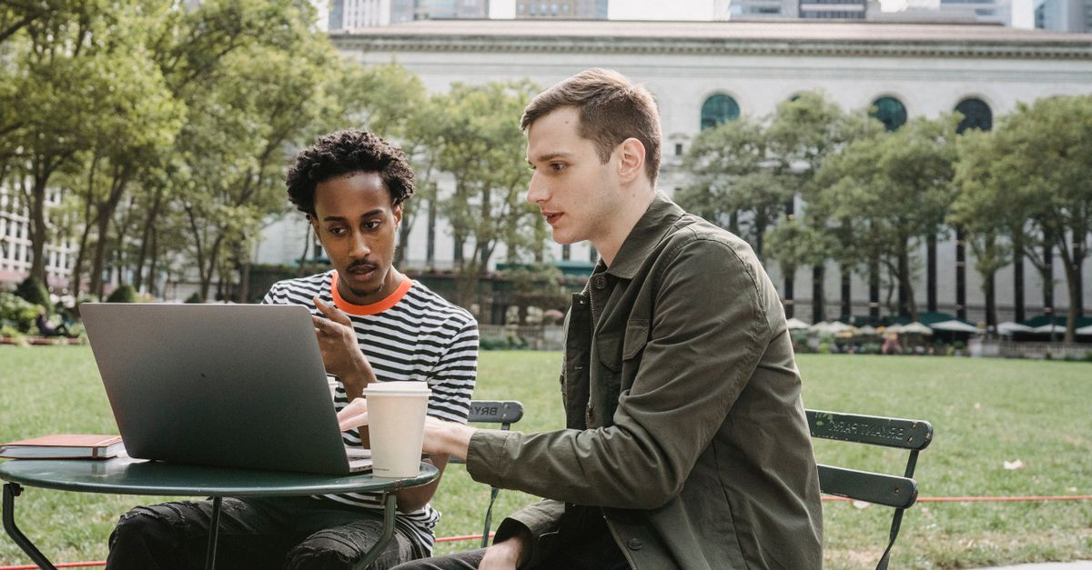 Focused young multiethnic male colleagues in casual clothes sitting at table in city park and working remotely on laptop