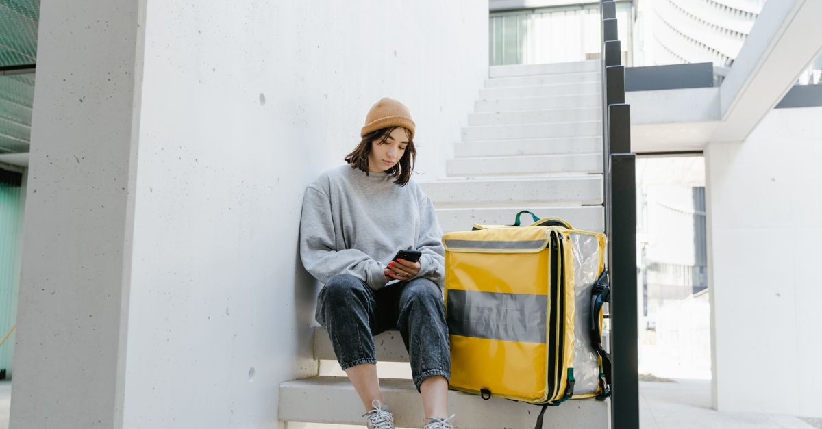 Woman courier in casual wear with a yellow thermal bag sitting on outdoor steps checking her phone.