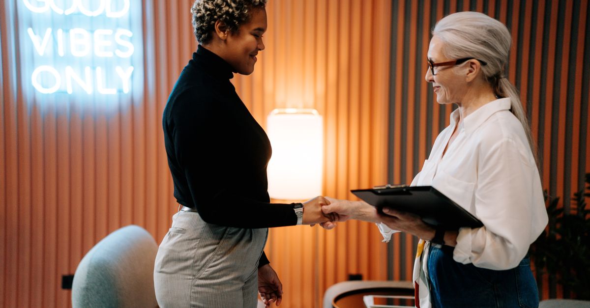 Two diverse businesswomen in a stylish office shaking hands under a 'Good Vibes Only' sign.