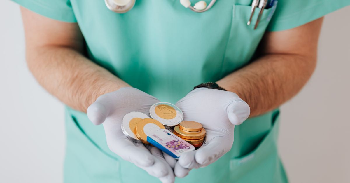 Unrecognizable medical worker demonstrating money wearing medicine form standing near white wall in hospital while working in day of salary