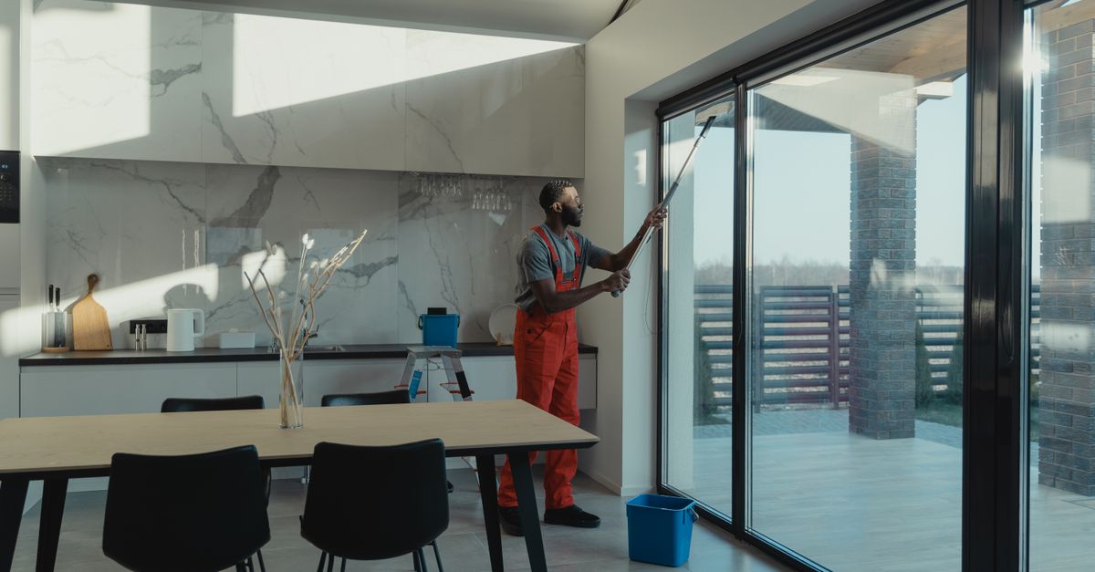 A professional cleaner in uniform washing large glass windows indoors with natural light streaming in.