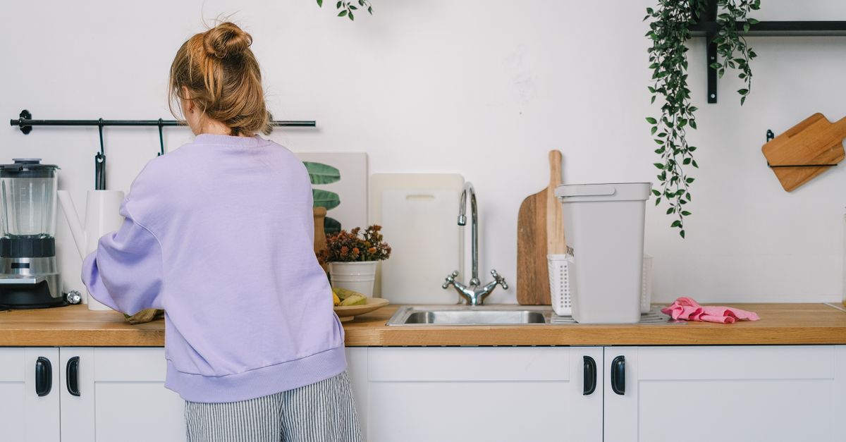 Back view of unrecognizable young female in casual clothes standing at wooden counter in light kitchen in daytime