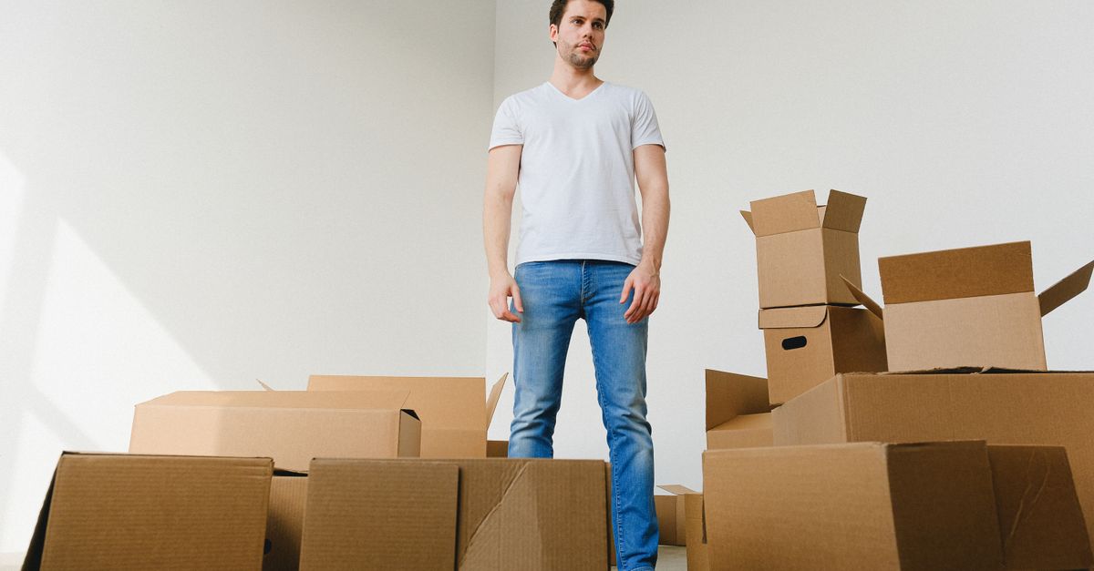 Low angle of serious young guy with dark hair in casual outfit standing near pile of cardboard boxes in light room and looking away while preparing for relocation