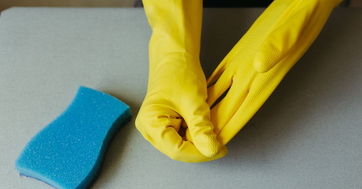 Close-up of hands wearing yellow gloves next to blue sponge on gray surface.