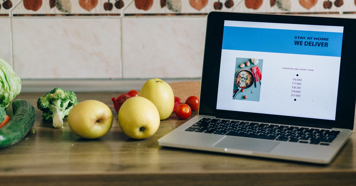Laptop showing a delivery app beside fresh fruits and vegetables on a kitchen counter.