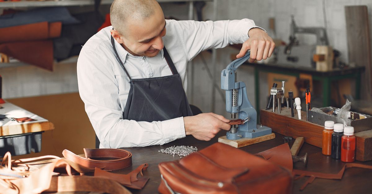 Happy young talented artisan in white shirt and apron attaching metal snaps on brown leather strap using pressing machine while working in workshop