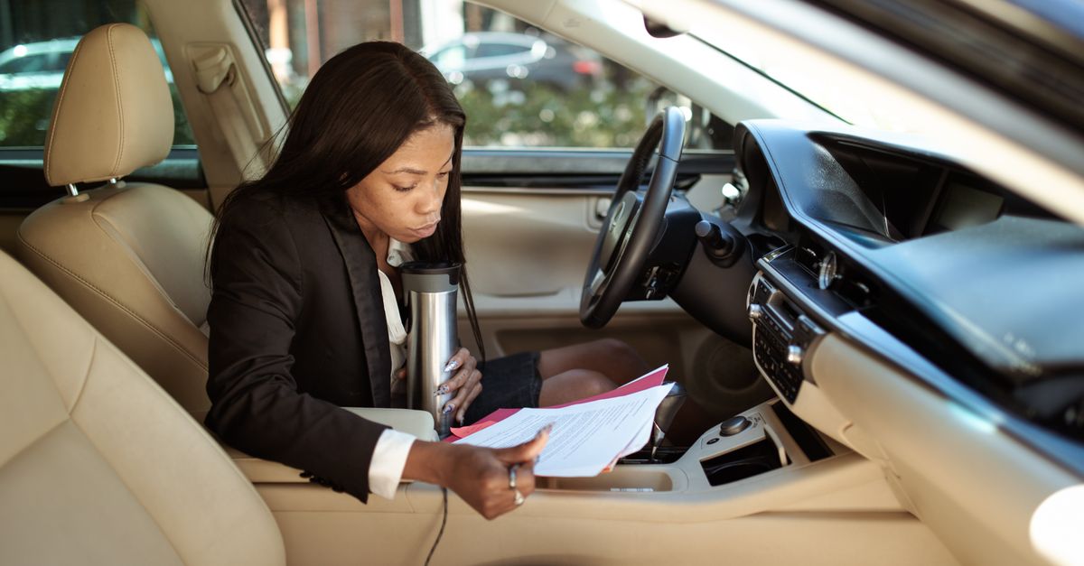 A focused businesswoman reads documents in her car, multitasking with a tumbler.