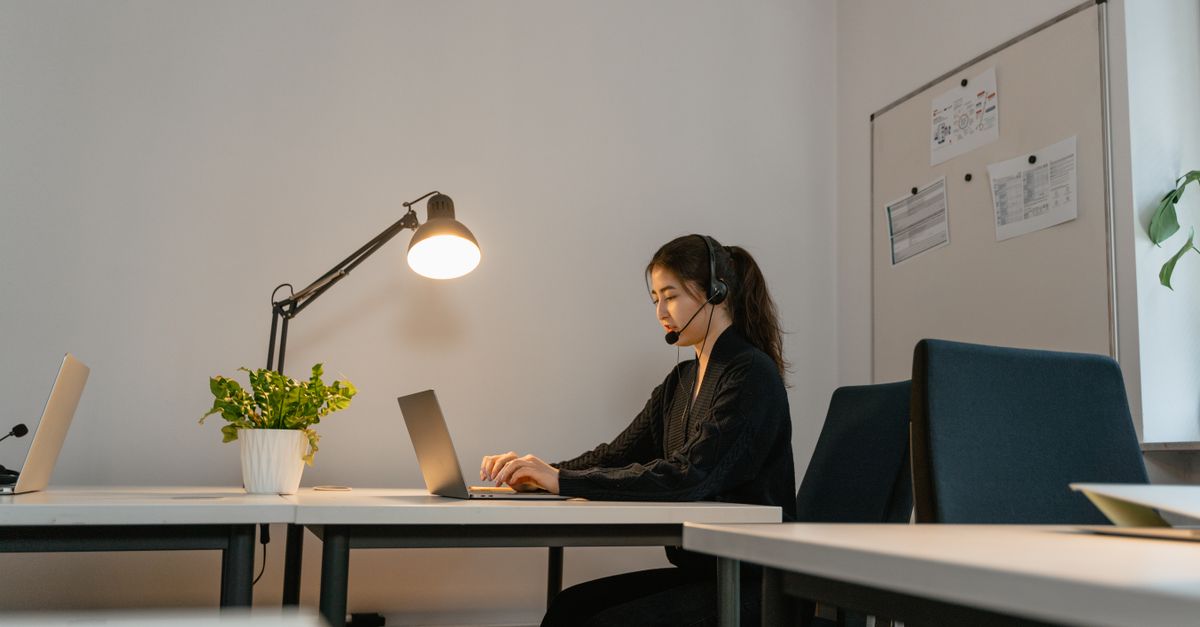 A woman in a modern office setting, wearing a headset and working on a laptop under a desk lamp.