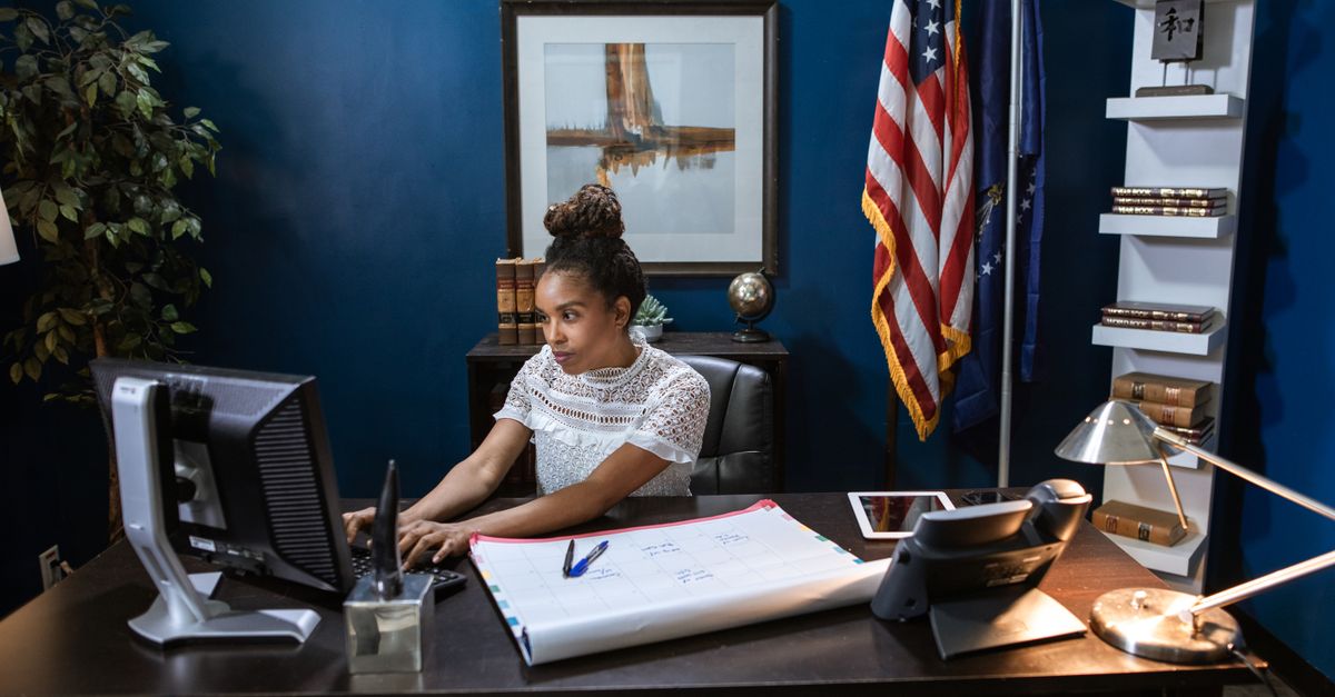 An African American woman concentrating on her work at a desk with computer and documents.