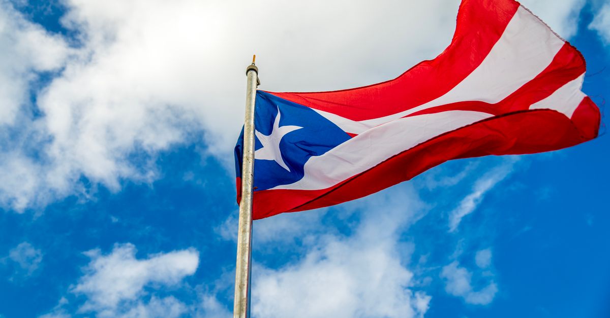 A vibrant Puerto Rican flag waving on a flagpole against a bright blue sky with clouds.
