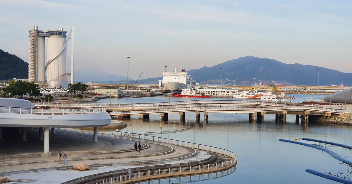 A contemporary harbor scene featuring ferries and architectural structures with a mountain backdrop under a clear sky.