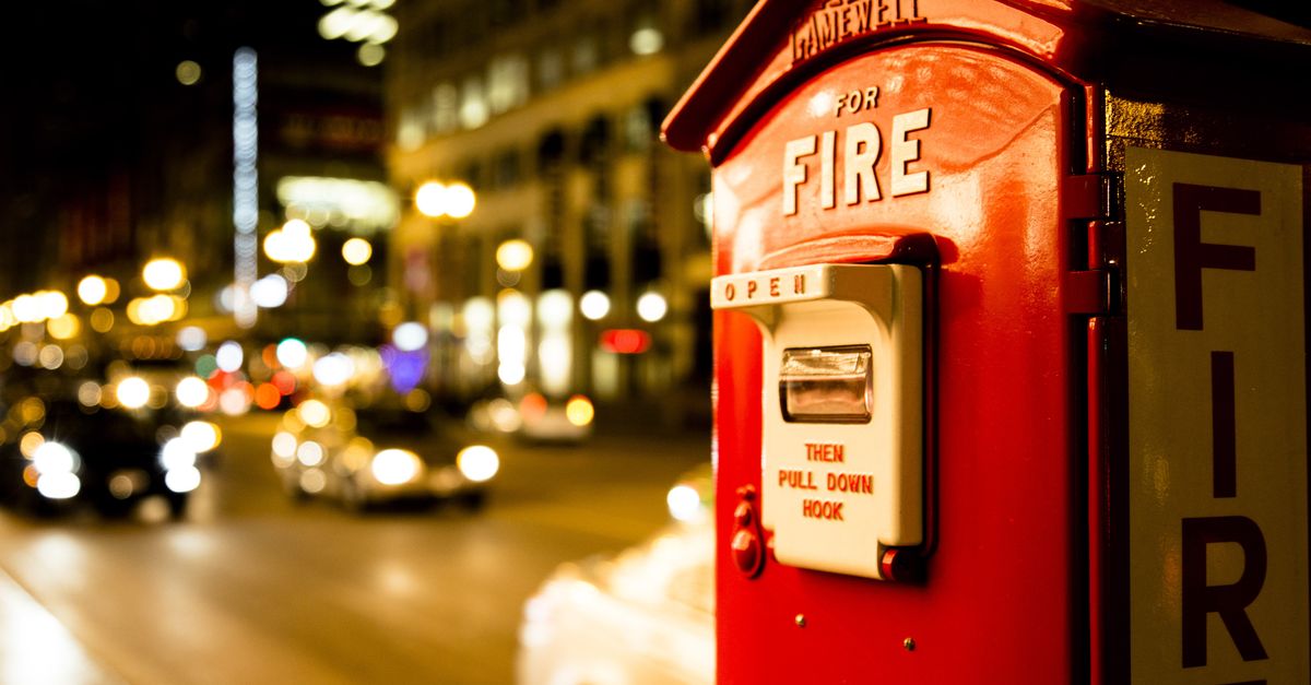 A classic red fire alarm box on a city street lit by car headlights at night.