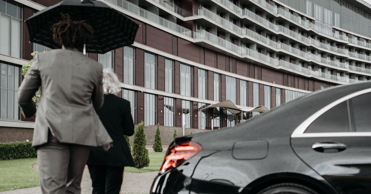 Two professionals in suits under an umbrella walking towards a modern building beside a luxury car.