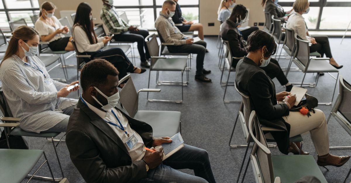 Business seminar with diverse attendees wearing masks, maintaining social distance indoors.