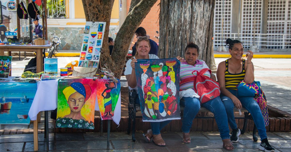 Three women sit at a colorful market stall displaying handmade paintings in an urban setting.