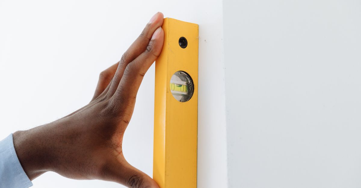 A close-up of a hand using a yellow level tool against a white wall to ensure vertical alignment.