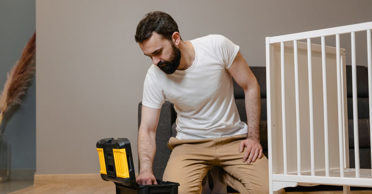 A man kneels indoors assembling a baby crib using a toolbox, wearing a white shirt and beige pants.