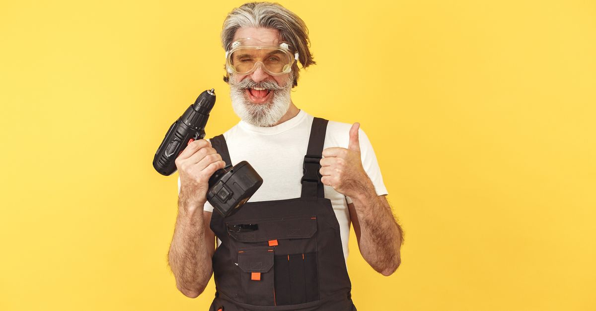 Senior man in safety gear and overalls holding a drill, smiling against a yellow background.