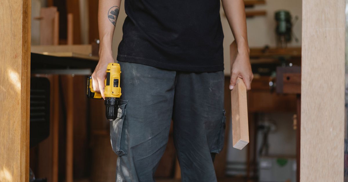Unrecognizable craftsman in casual clothes standing with electric screwdriver and piece of wood while working in professional workshop