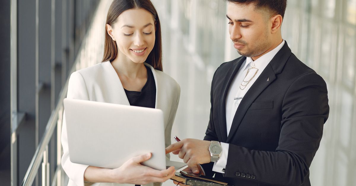 Concentrated diverse male and female employees in formal clothes standing in modern office corridor and browsing laptop while discussing business project together