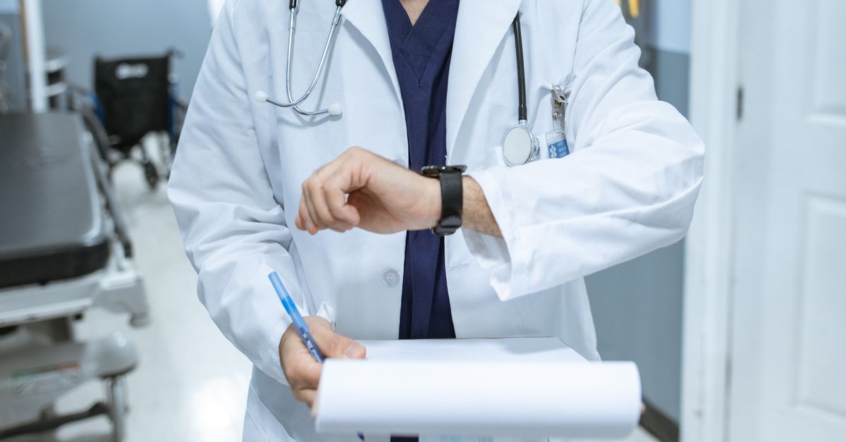 Doctor in lab coat checking wristwatch while holding medical chart in hospital corridor.