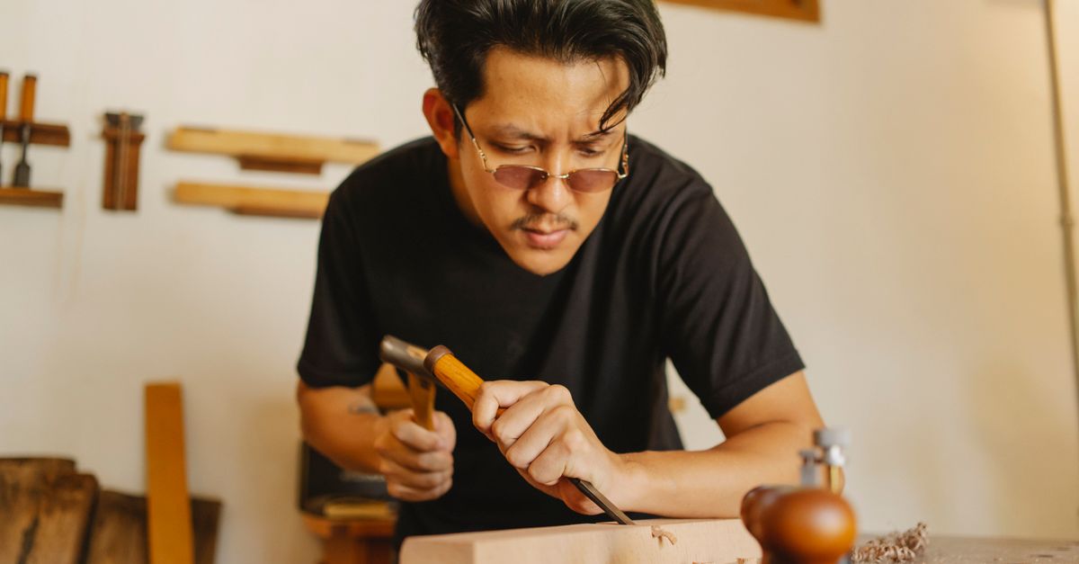 Concentrated Asian man working with chisel and hammer while cutting notch in hardwood plank