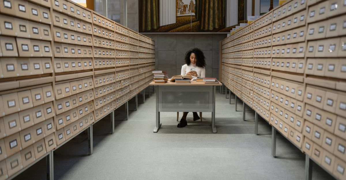 A young woman reading at a desk in an organized library archive room with wooden drawers.
