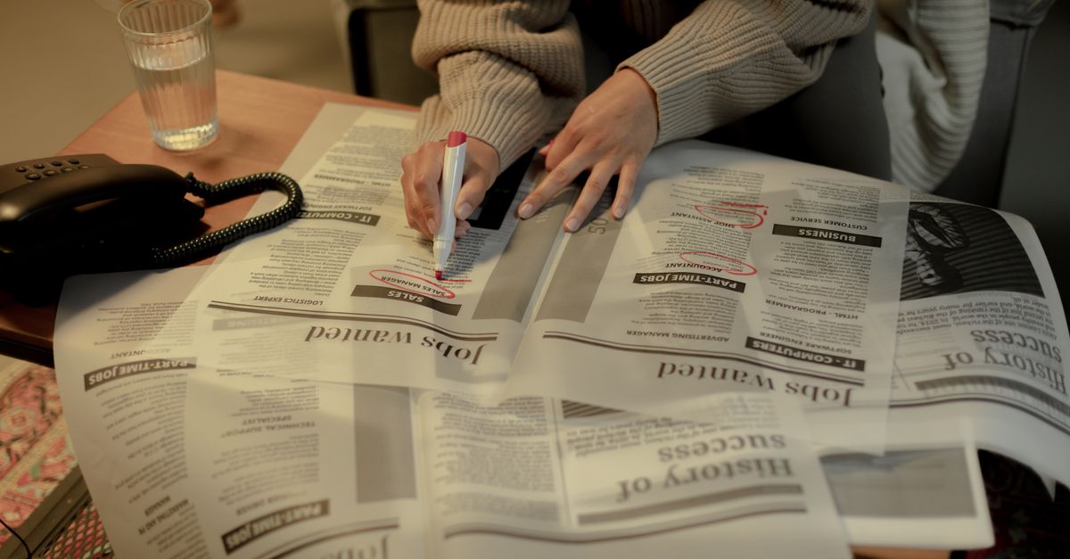 An adult woman marks job listings in a newspaper while sitting indoors, highlighting job search activity.