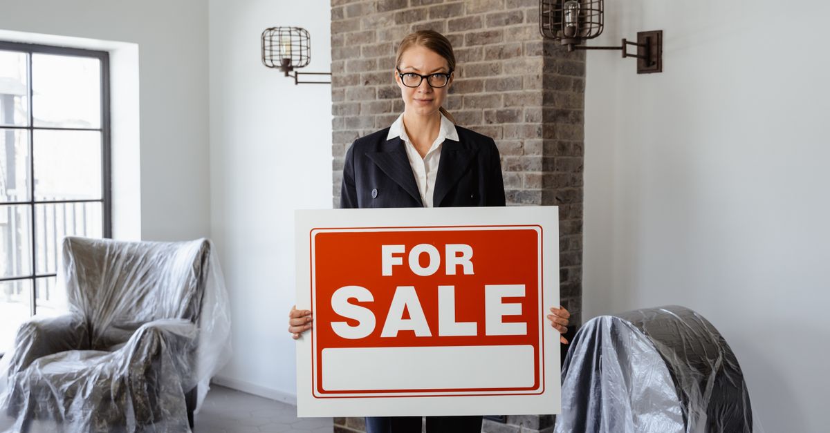 Real estate agent holding a 'For Sale' sign in a furnished living room.