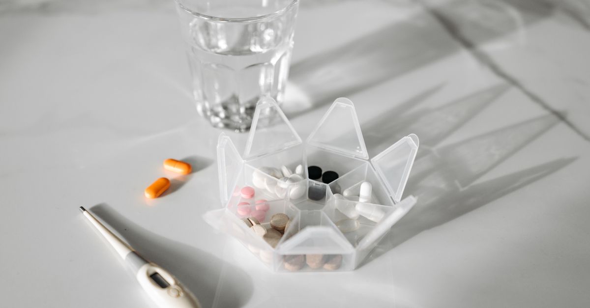 A pill organizer with medication, a glass of water, and a thermometer on a white surface.