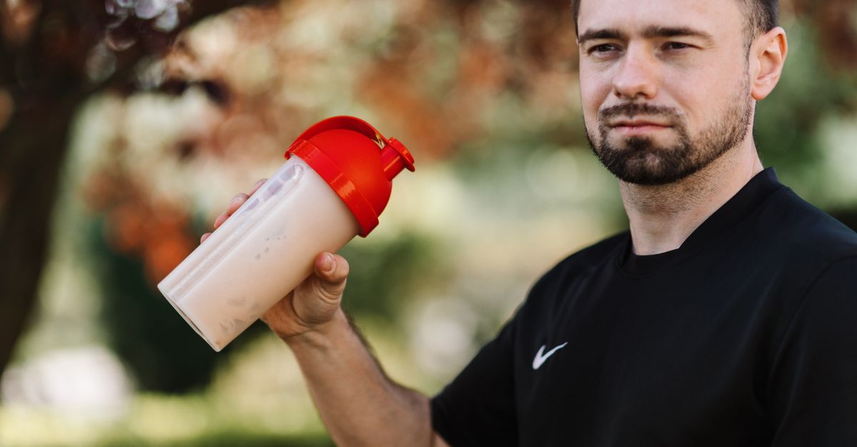 Adult man holds a protein shake in a red lid tumbler outdoors, wearing a black shirt.