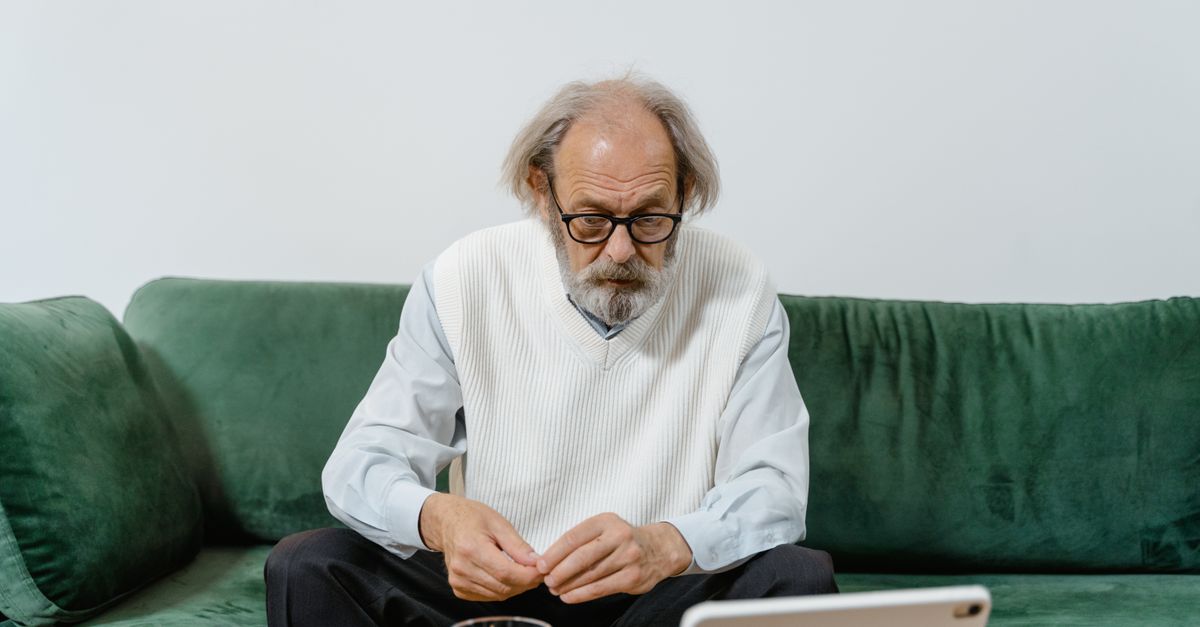 Senior man sitting on green sofa, focusing on a tablet, possibly for telemedicine or social interactions.