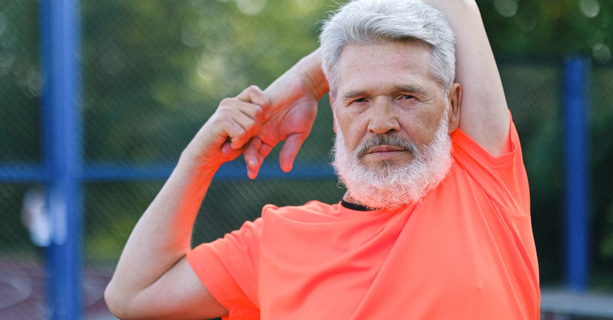 Confident bearded male with gray hair in sportswear stretching arms during workout on sports ground and looking at camera