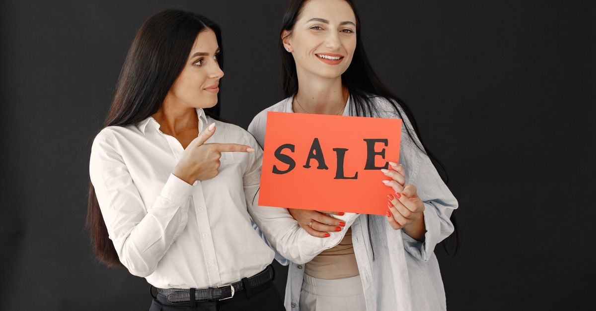 Two women with long hair smiling and holding a sale sign against a black background.