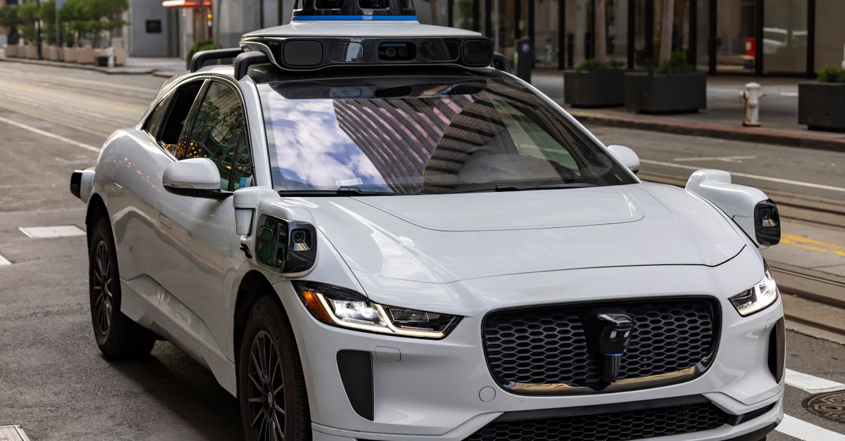 A white autonomous vehicle navigating a city street, reflecting urban architecture in daylight.