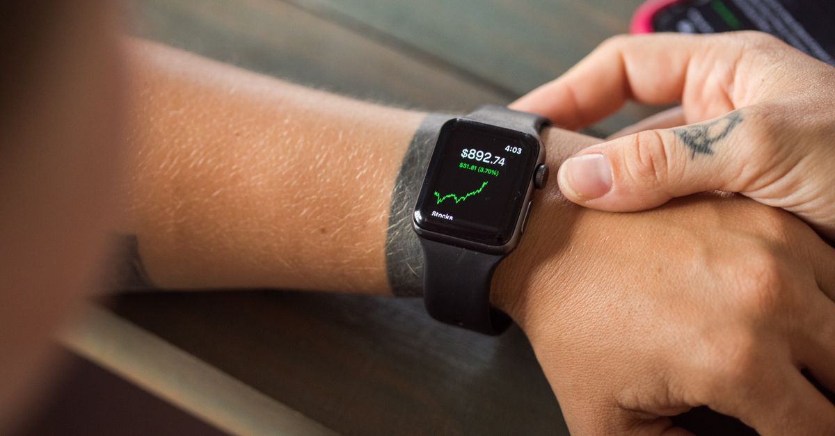Close-up of a smartwatch showing stock market data, with hands wearing it.