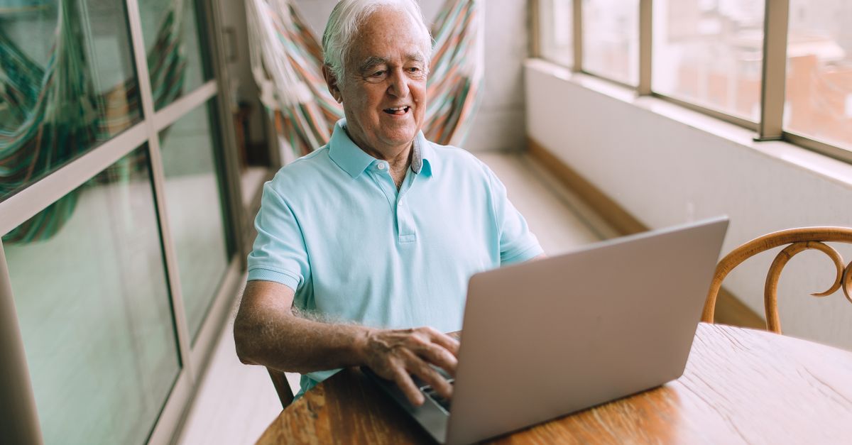 Elderly man happily using a laptop on a sunny balcony, embracing online connectivity.