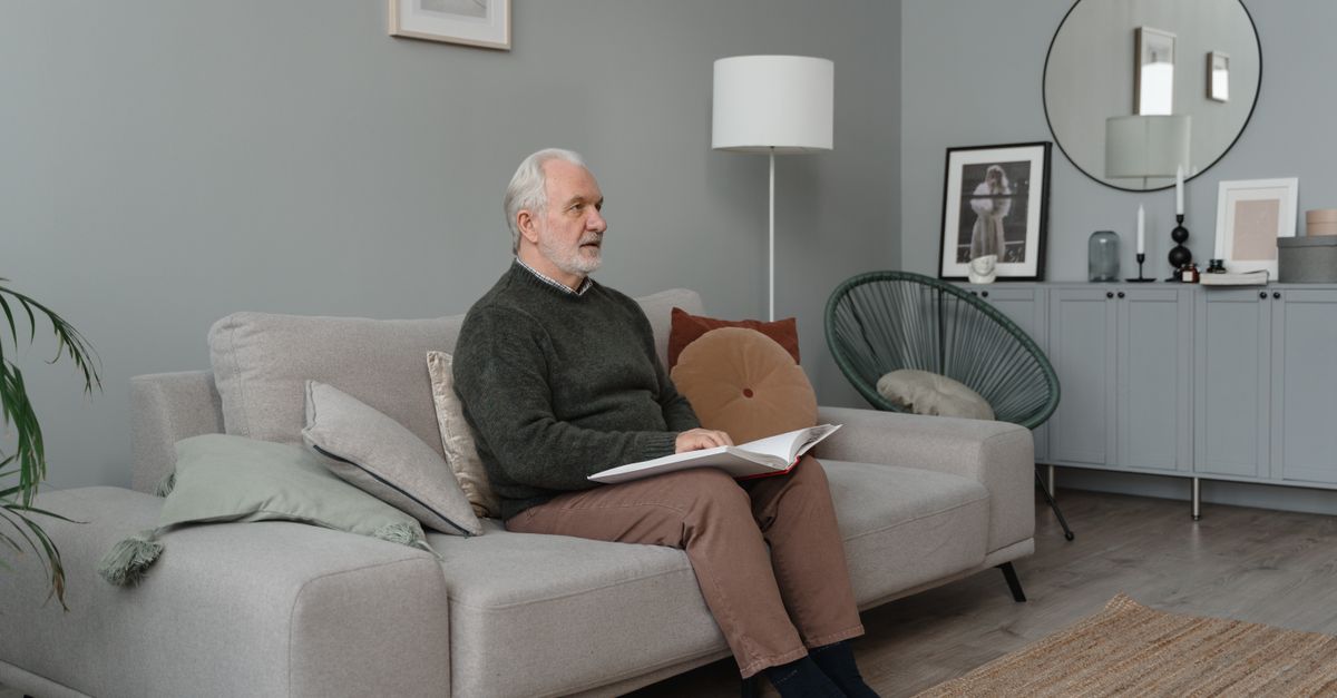 Elderly man reading a Braille book while sitting on a comfortable sofa in a modern living room.