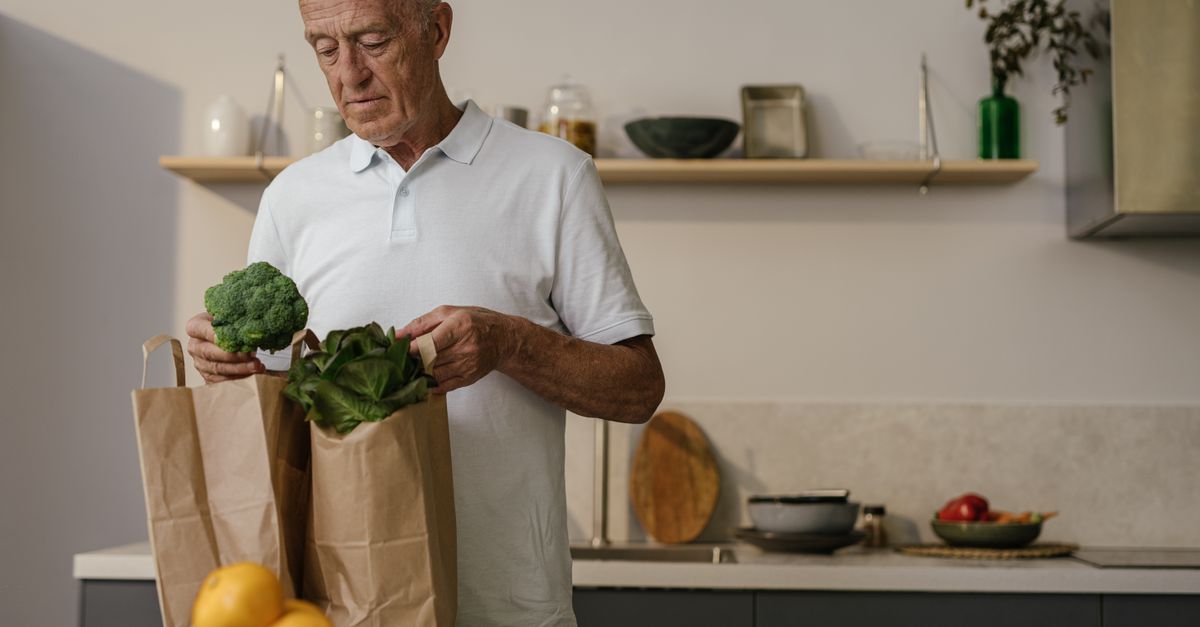 Elderly man in kitchen examining fresh vegetables from paper bags. Healthy lifestyle concept.